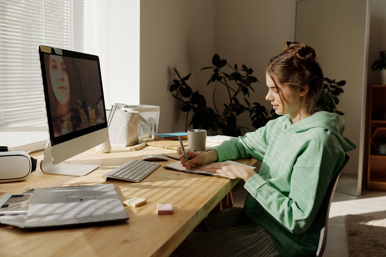 Teenager in hoodie focused on remote learning via video call, taking notes at home desk.