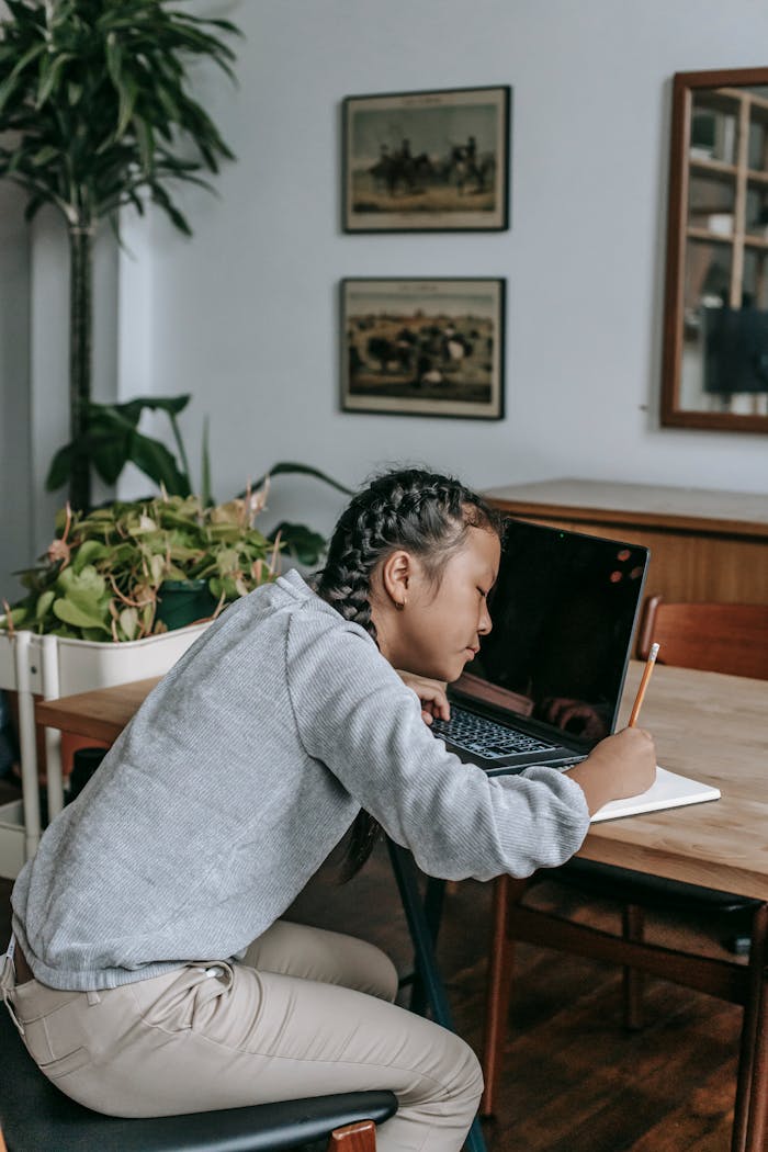 Home Side view of concentrated Asian girl in casual clothes sitting at table with opened laptop and writing in copybook