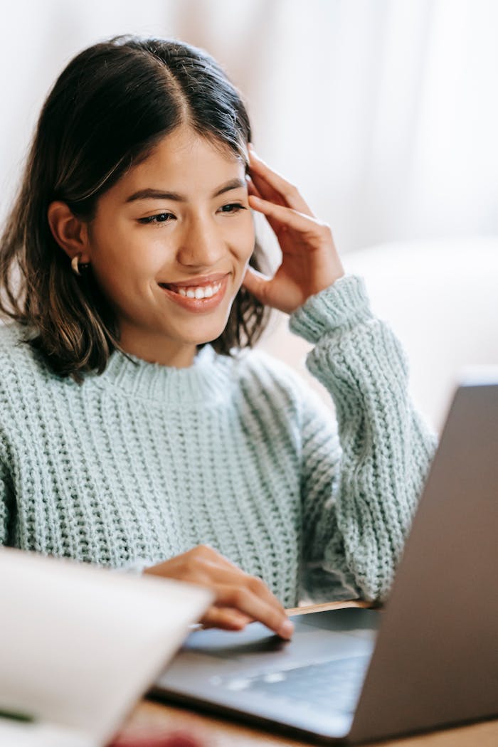 Smiling young Latin American female freelancer in casual clothes at table browsing on netbook near notebook with pen in light room