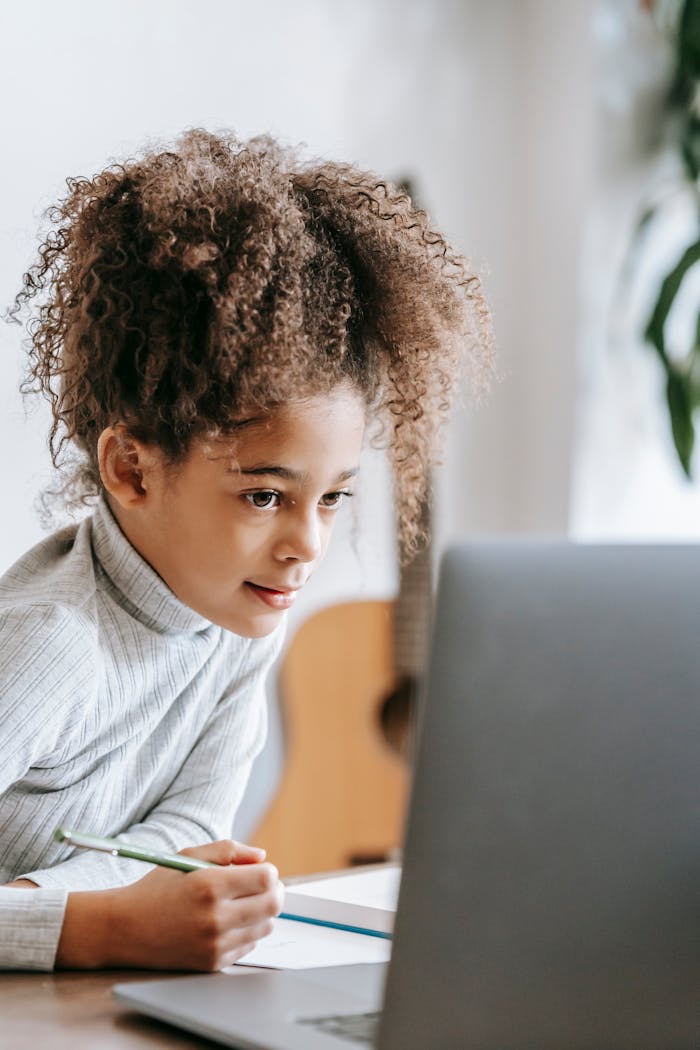 Home Positive African American girl with curly hair taking notes on paper while looking at screen of netbook during studies at home with guitar on blurred background