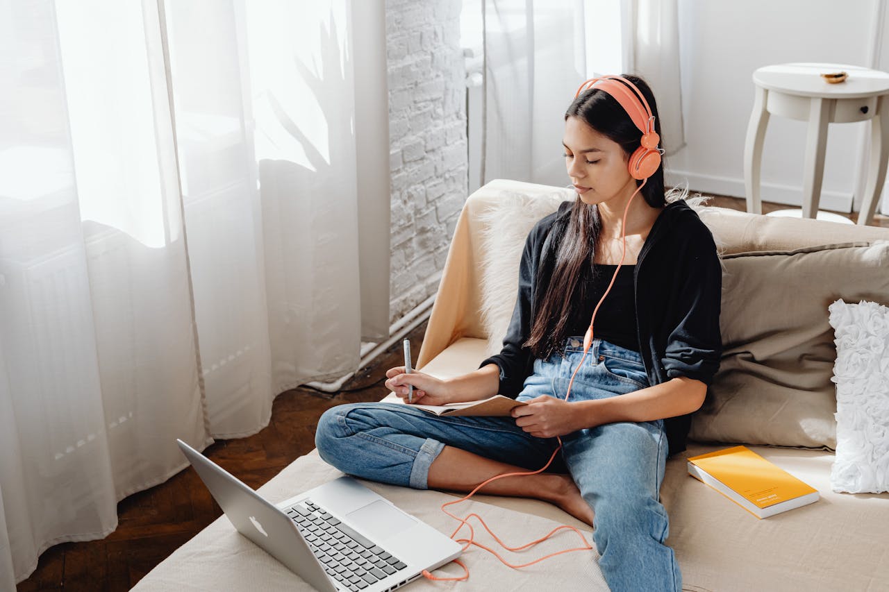 Young woman with headphones studying online from home on a sofa using a laptop.