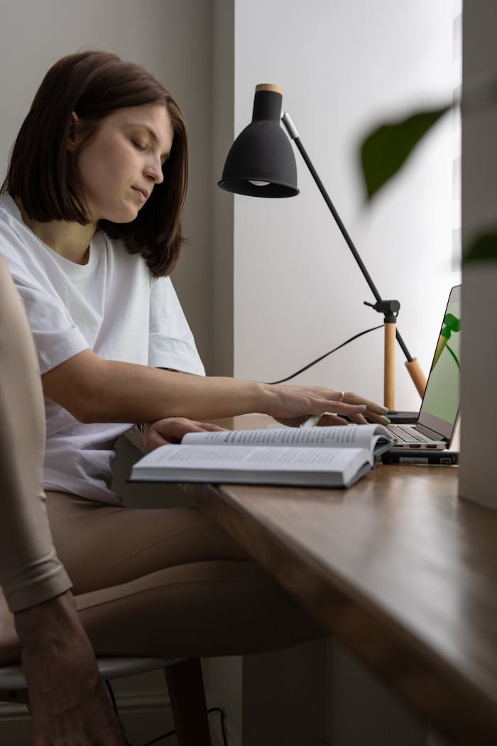 Side view of concentrated female student reading book while surfing internet on netbook at wooden table with lamp in room