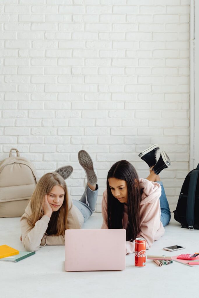 Two teenage girls lying on the floor studying with a laptop and books.