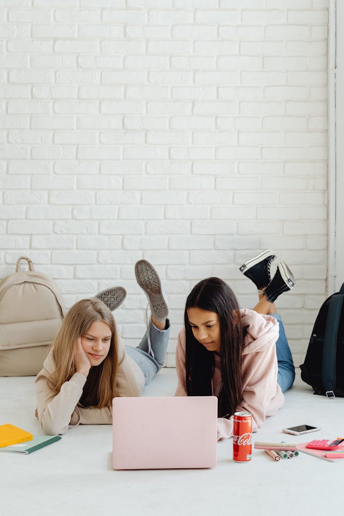 Two teenage girls lying on the floor studying with a laptop and books.