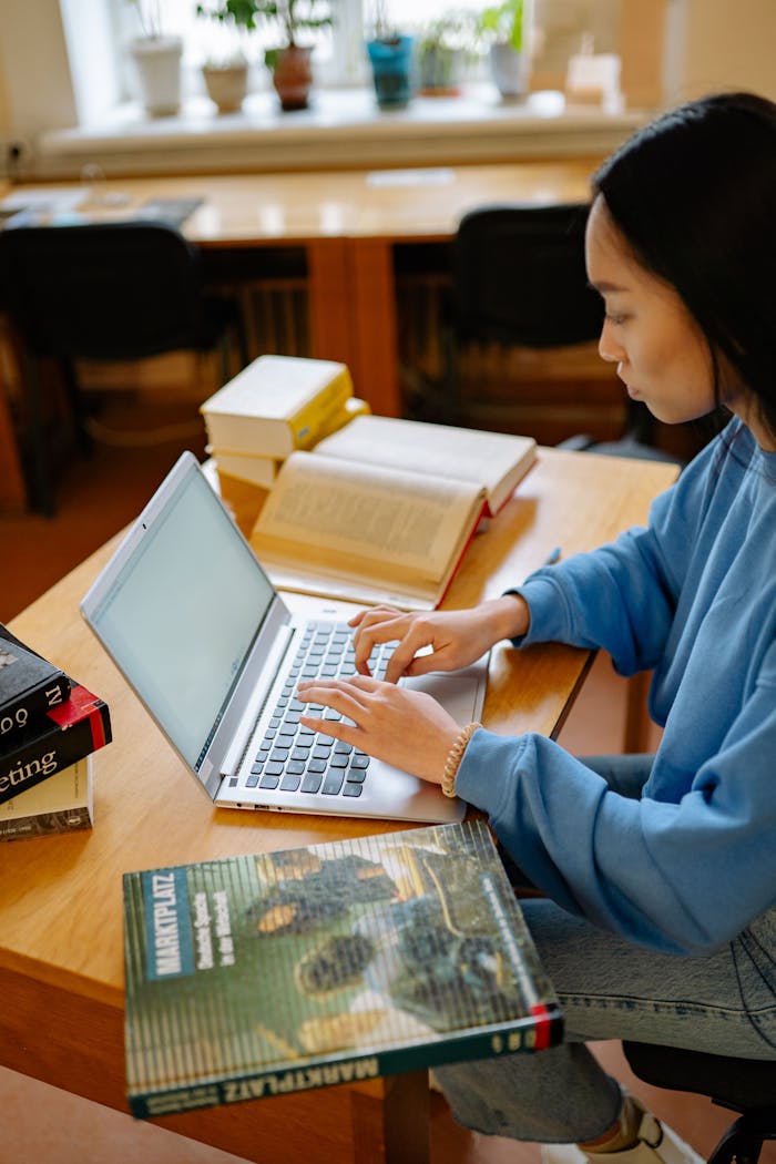 Home A young woman is focused on studying with her laptop at a wooden desk with open books.